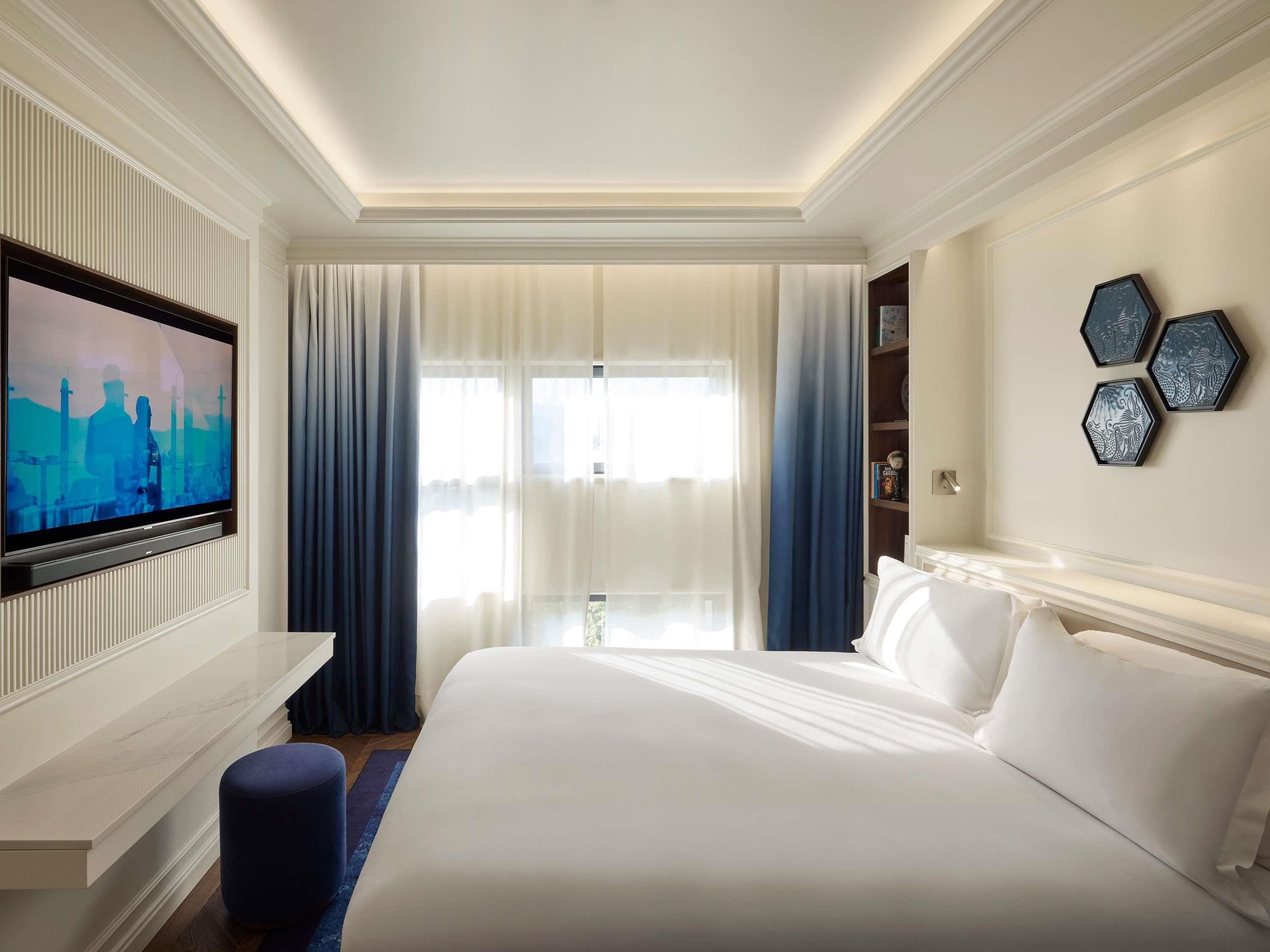 Quarto de hotel com uma cama grande, televisão de tela plana na parede, estante de livros e janela com cortinas.