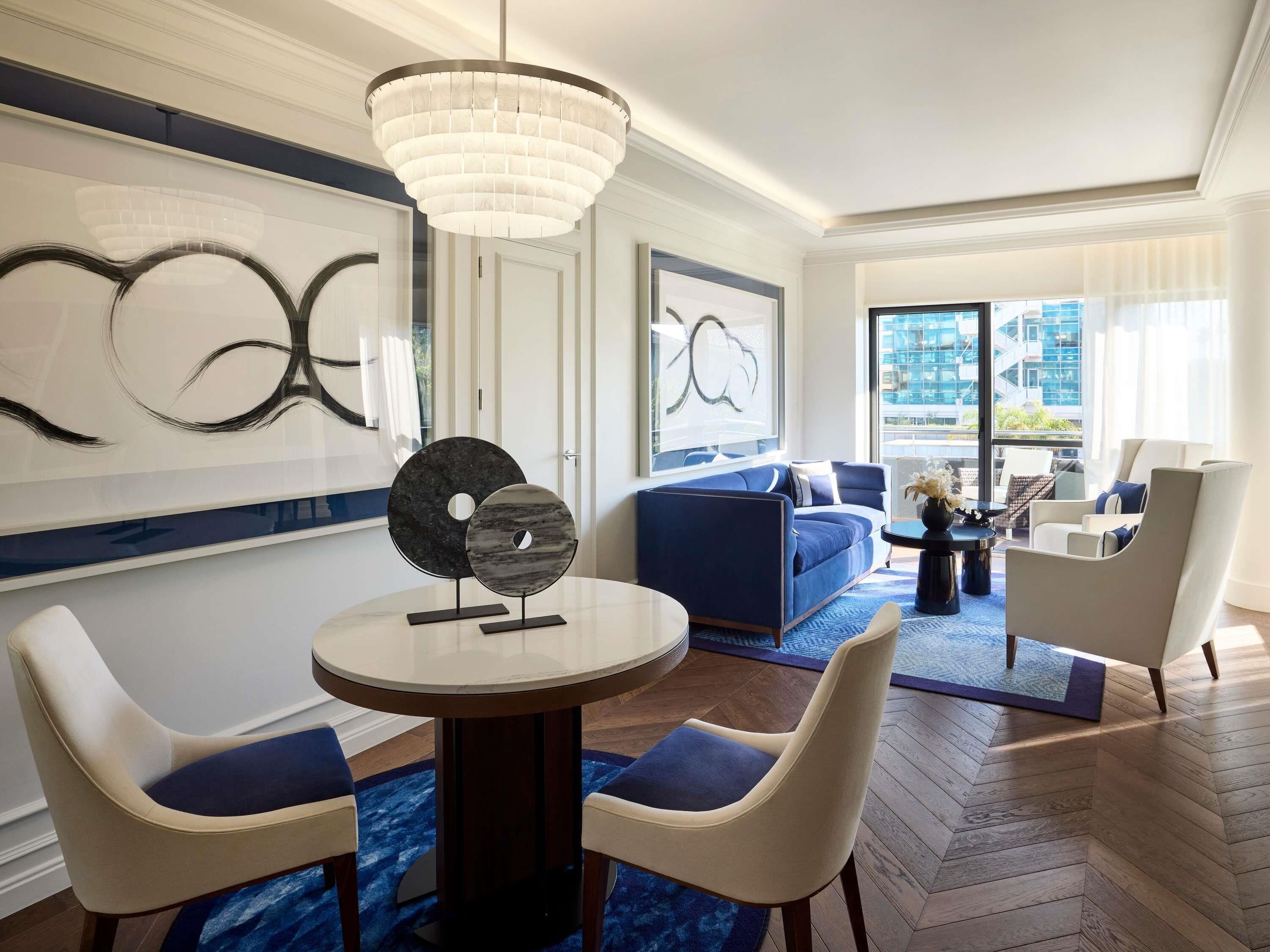 A modern hotel room with a round dining table, two chairs, a blue sofa, a coffee table, and a large window with a city view.
