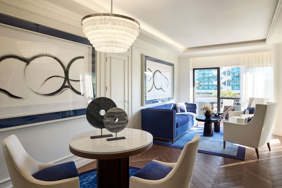 Ein modernes Hotelzimmer mit einem runden Esstisch, zwei Stühlen, einem blauen Sofa, einem Couchtisch und einem großen Fenster mit Blick auf die Stadt.