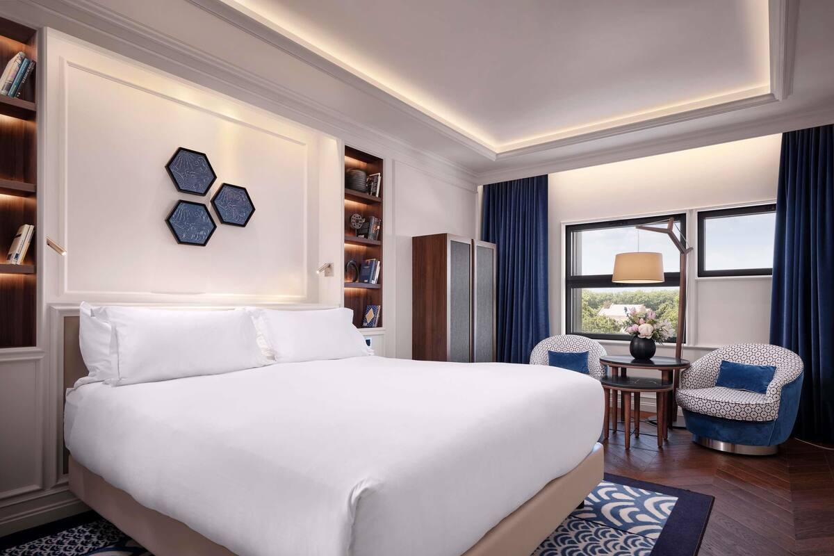 Ein Hotelzimmer mit einem großen Bett, einem Schreibtisch mit Stuhl, einem Bücherregal und einem Fenster mit Vorhängen.