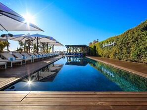 Zona de piscina con sillones de descanso y sombrillas, una terraza de madera y cielo despejado.