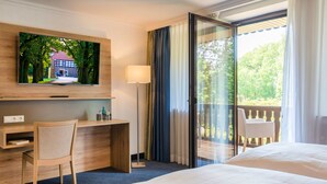 A hotel room with a bed, a desk with a chair, a TV mounted on the wall, and a balcony with a view of trees and a building.