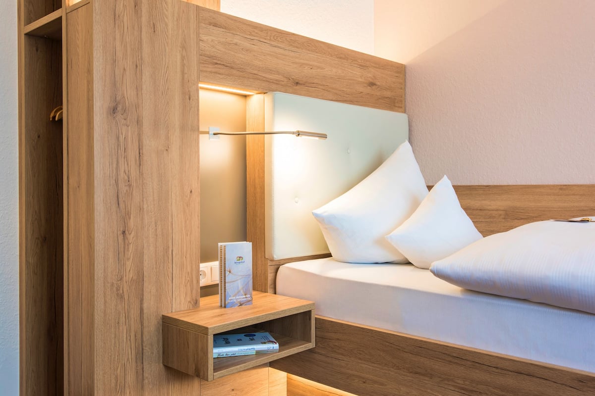 Une chambre d’hôtel moderne avec une tête de lit en bois, une table de chevet avec une lampe et un lit bien fait, recouvert de draps blancs.