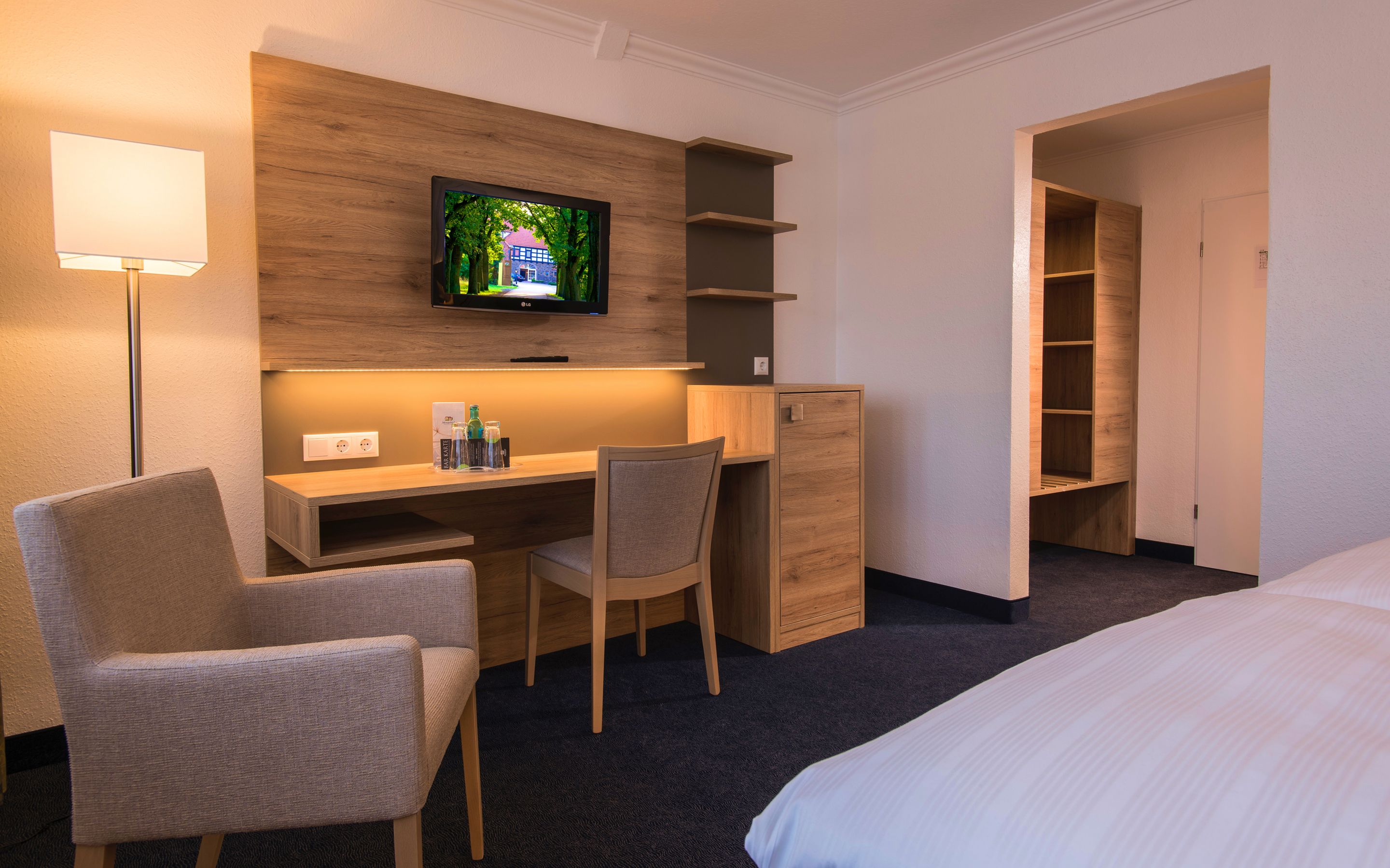 A hotel room with a bed, a desk with a chair, a TV mounted on the wall, and a wardrobe.