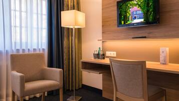 Ein Hotelzimmer mit einem wandmontierten Flachbildfernseher, einem Schreibtisch mit Lampe, zwei Stühlen und Blick auf ein Gebäude durch das Fenster.