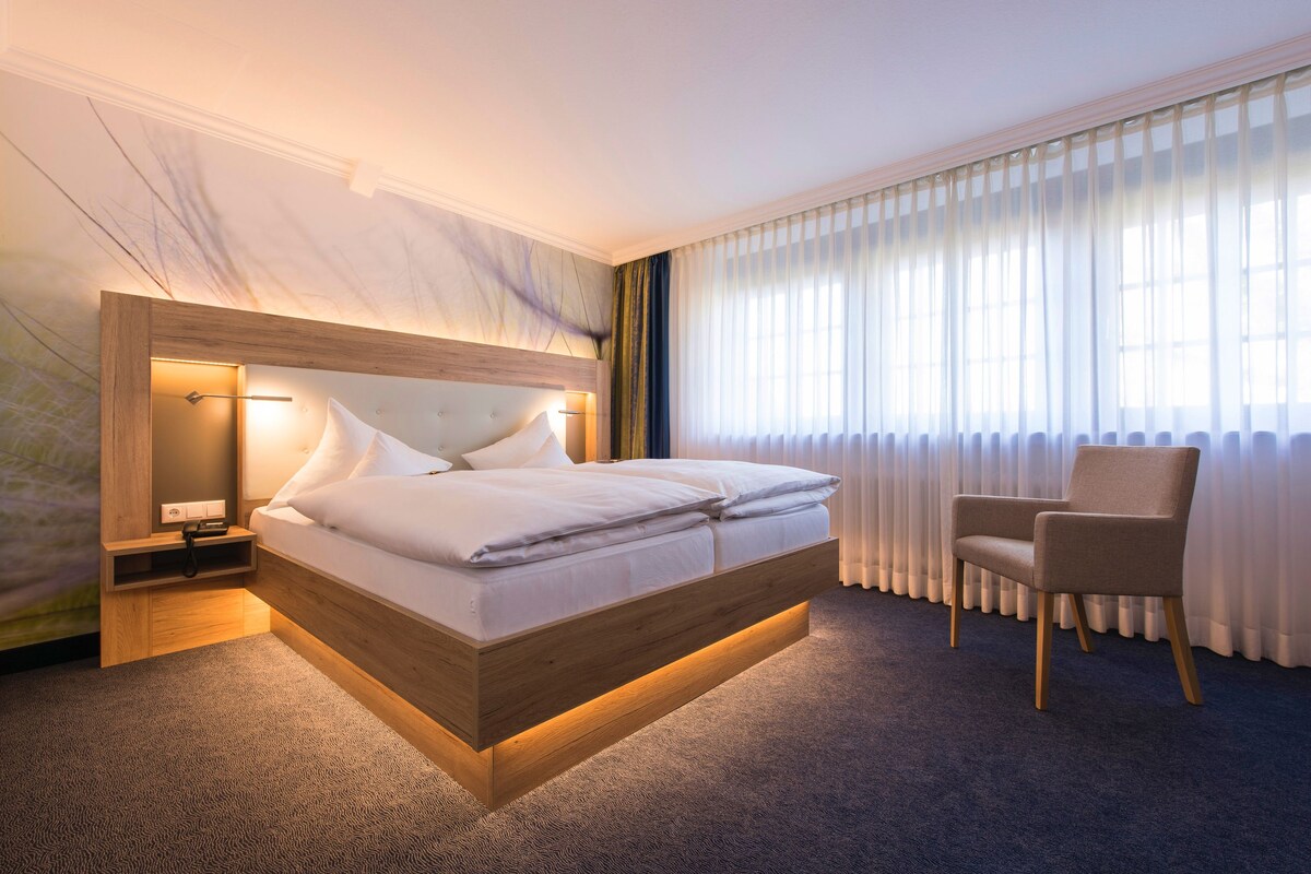 A modern hotel room with a wooden bed, a bedside table, a chair, and a large window with sheer curtains.