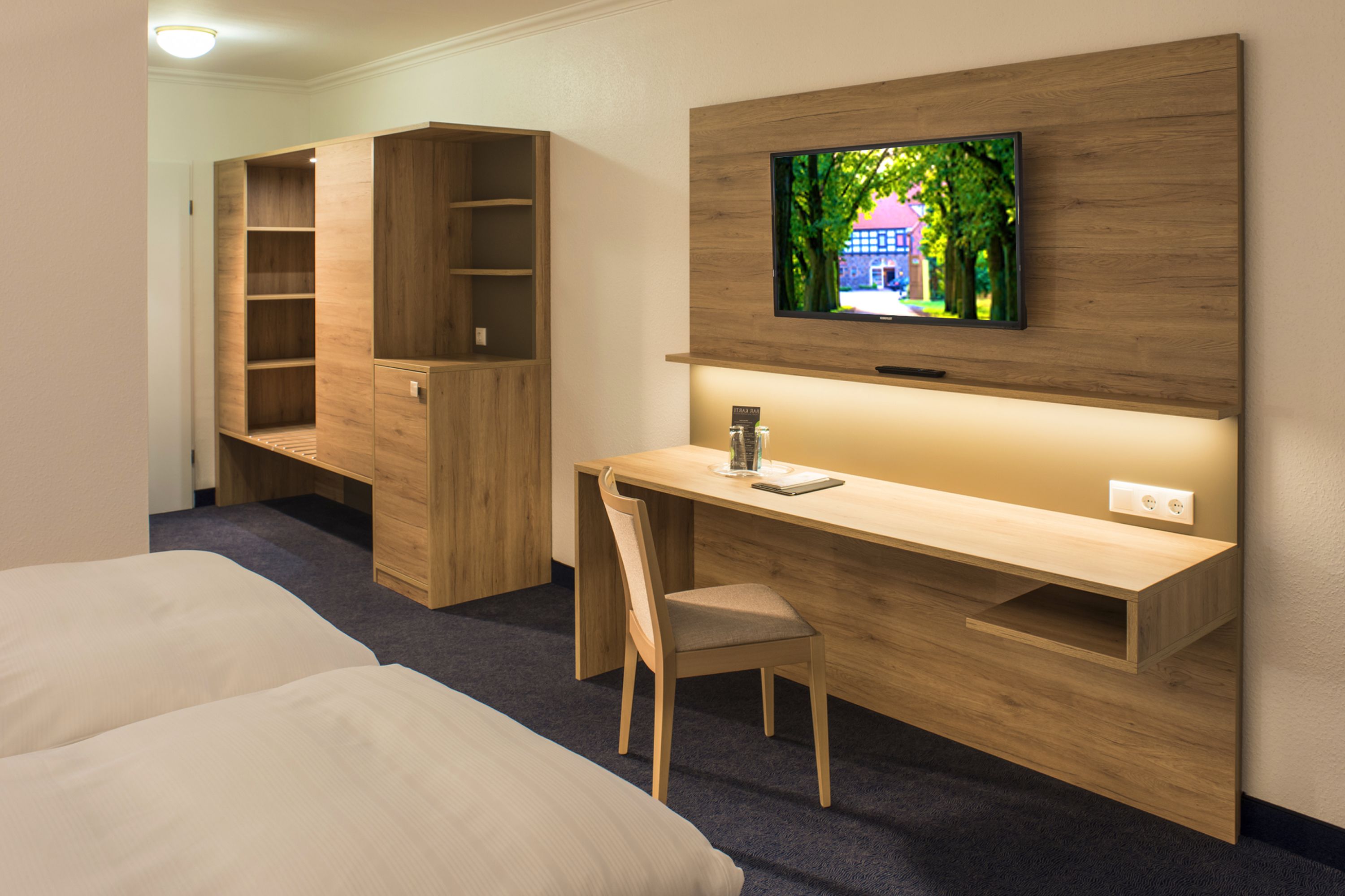 A hotel room with a wooden desk, a television mounted on the wall, a chair, and two beds.