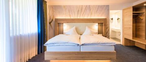 A modern hotel room with a wooden headboard, a large bed with white linens, a wooden wardrobe, and a bathroom with a wooden vanity.