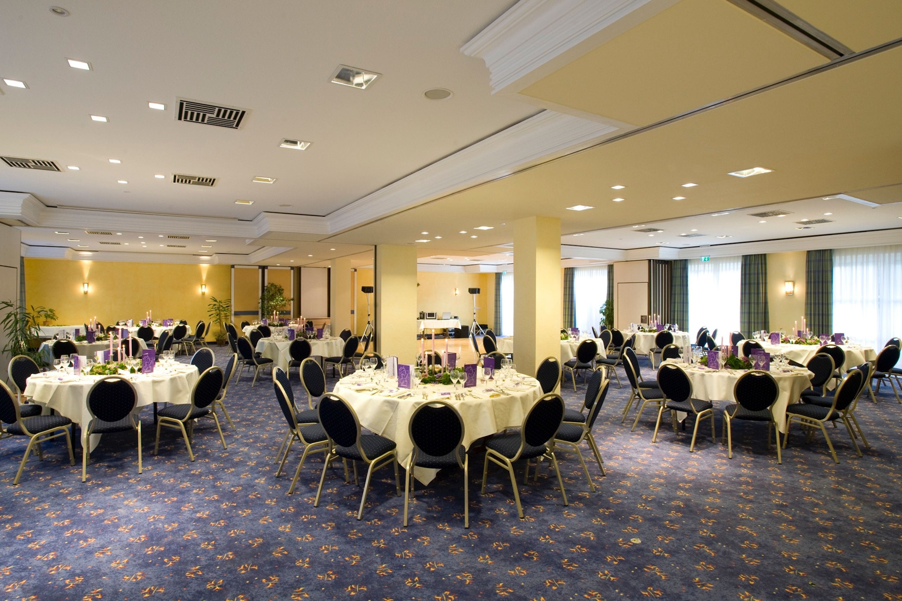 Ein großer Festsaal mit mehreren runden Tischen, gedeckt zum Essen, mit Teppichboden und Deckenbeleuchtung.