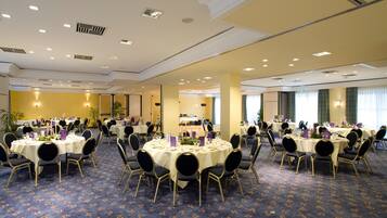 A large banquet hall with multiple round tables set for dining, featuring a carpeted floor and ceiling with recessed lighting.