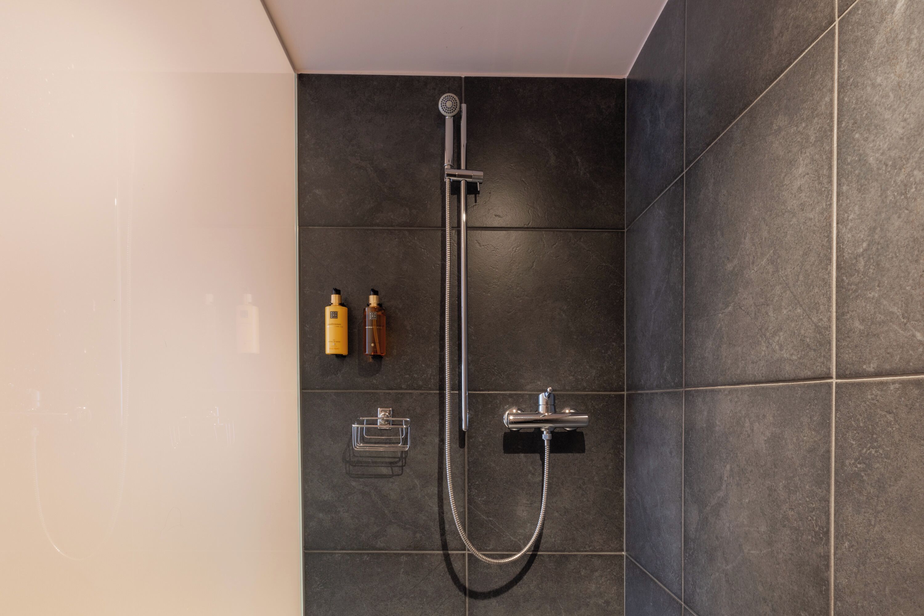 A modern shower with a handheld showerhead and a wall-mounted control. There are two bottles of shampoo and a soap dish on the wall.