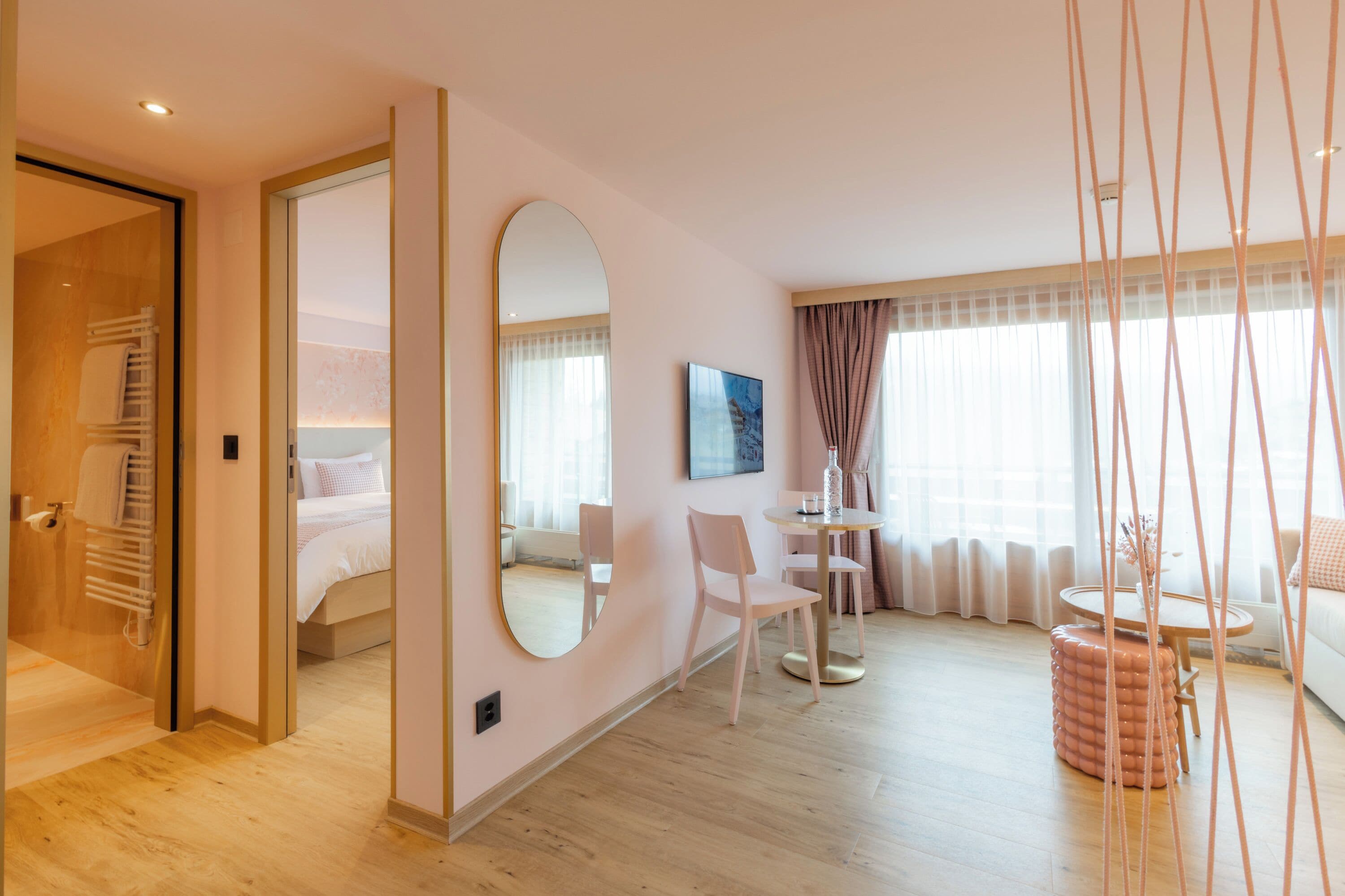 Una habitación de hotel moderna con piso de madera, una cama con ropa blanca, una mesa redonda con sillas, un otoman redonda y un espejo grande en la pared.