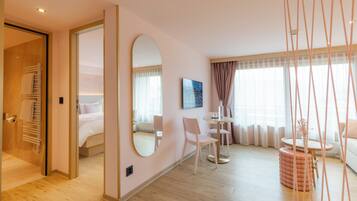 Una habitación de hotel moderna con piso de madera, una cama con ropa blanca, una mesa redonda con sillas, un otoman redonda y un espejo grande en la pared.