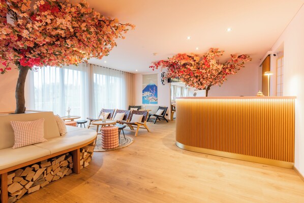 A modern hotel lobby with a wooden reception desk, a seating area with sofas and chairs, and a large tree with pink flowers.