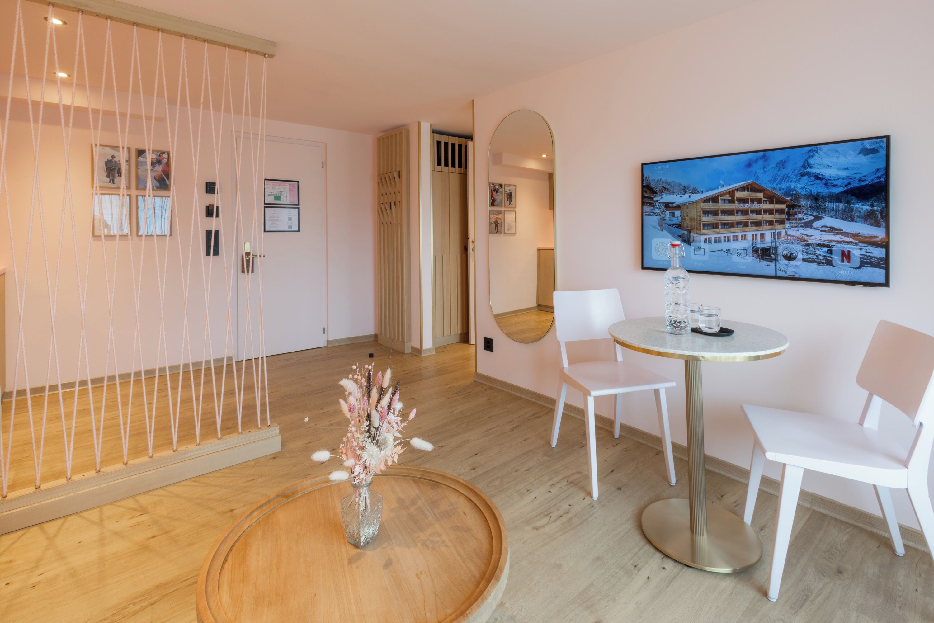A hotel room with a wooden floor, a round wooden table with a vase of flowers, two white chairs, a large framed picture of a mountain lodge, and a sliding door with a view of a balcony.