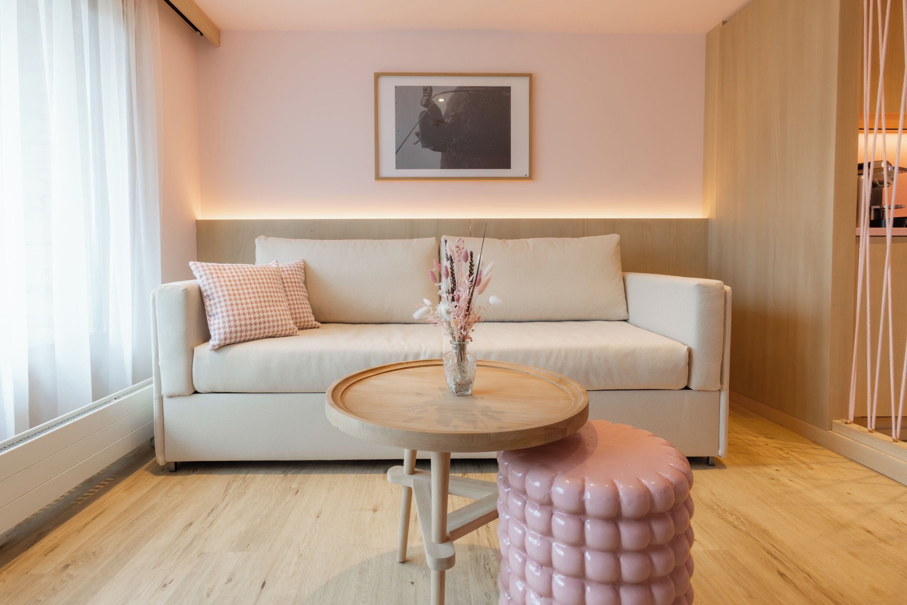 A modern living room with a sofa, a round wooden table, and a pink ottoman.