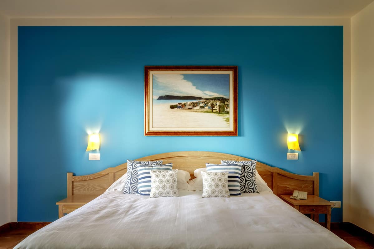 A hotel room with a wooden headboard, a bed with white bedding, two patterned pillows, a nightstand with a phone, and a framed picture on the wall.