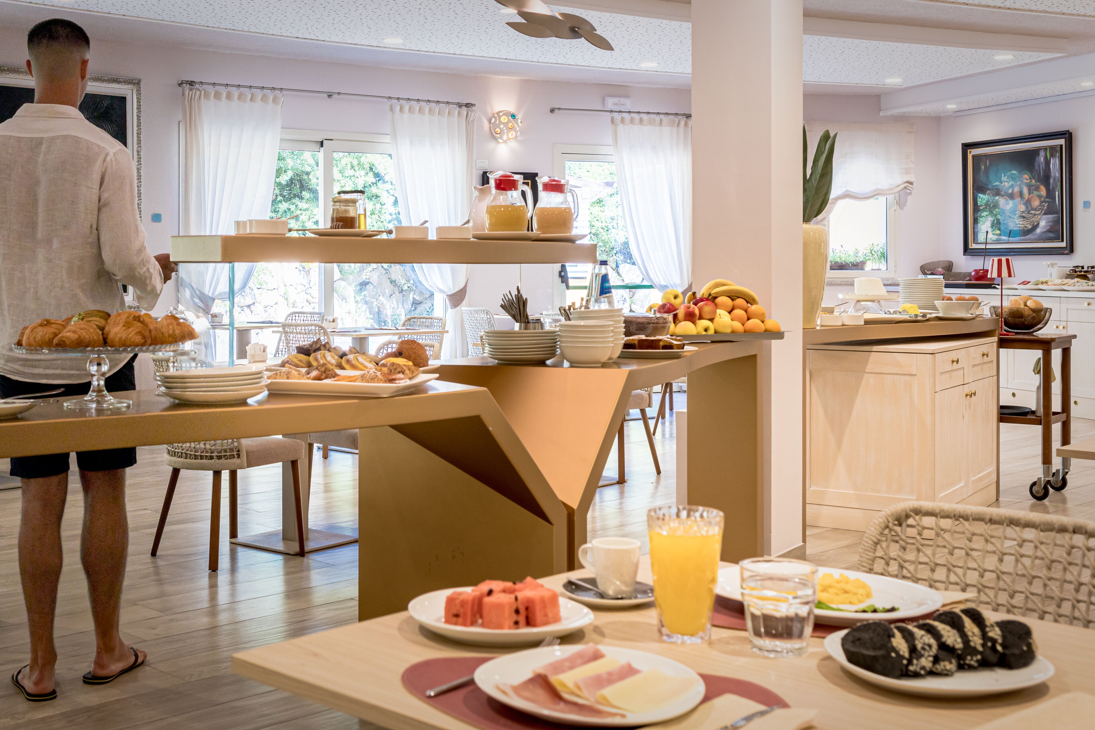 Un bufé de desayuno con diversos alimentos y bebidas, una persona de pie junto al bufé, y una zona de cocina al fondo.