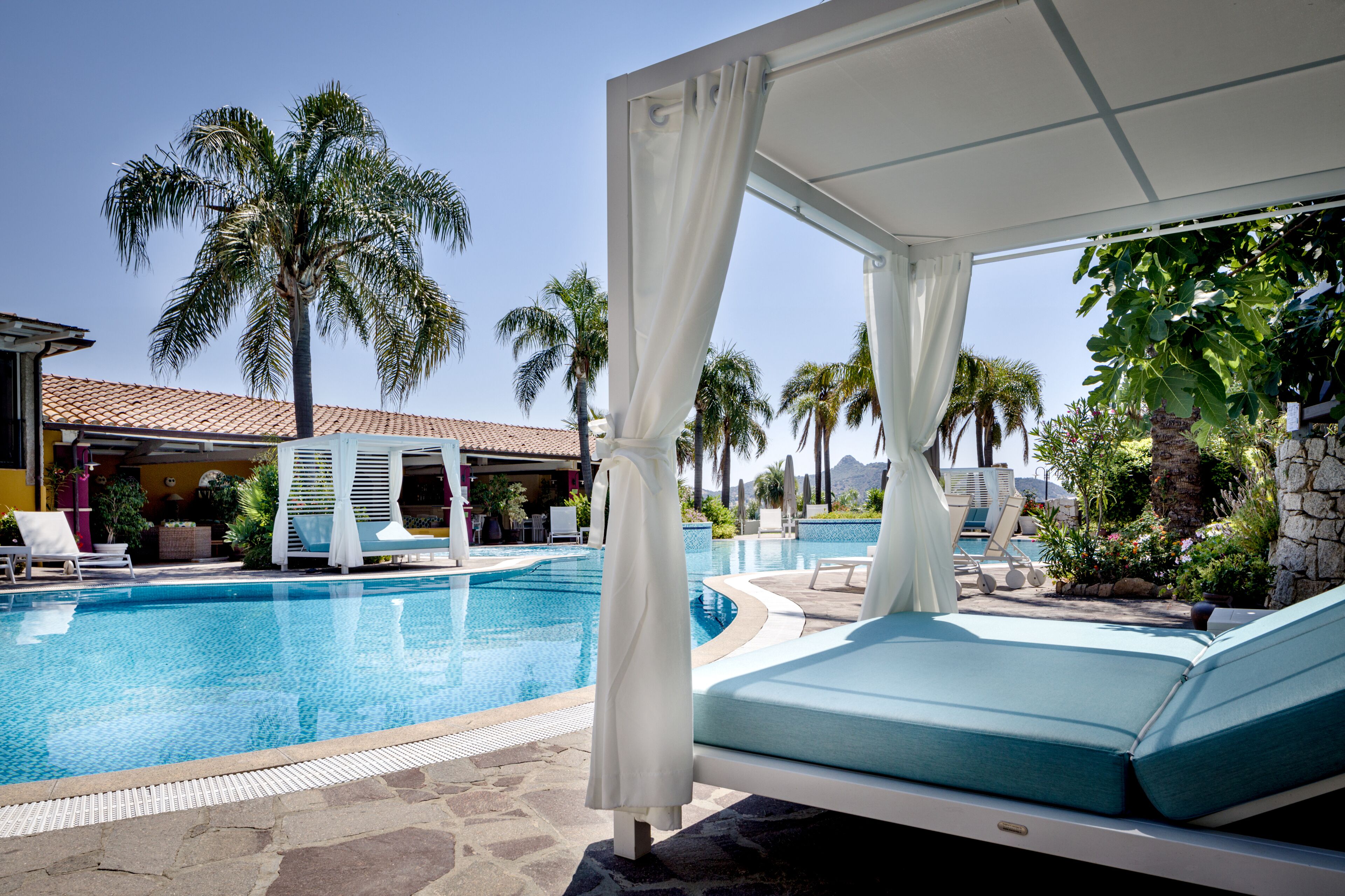 A pool area with a lounge chair, a covered seating area, and palm trees in the background.