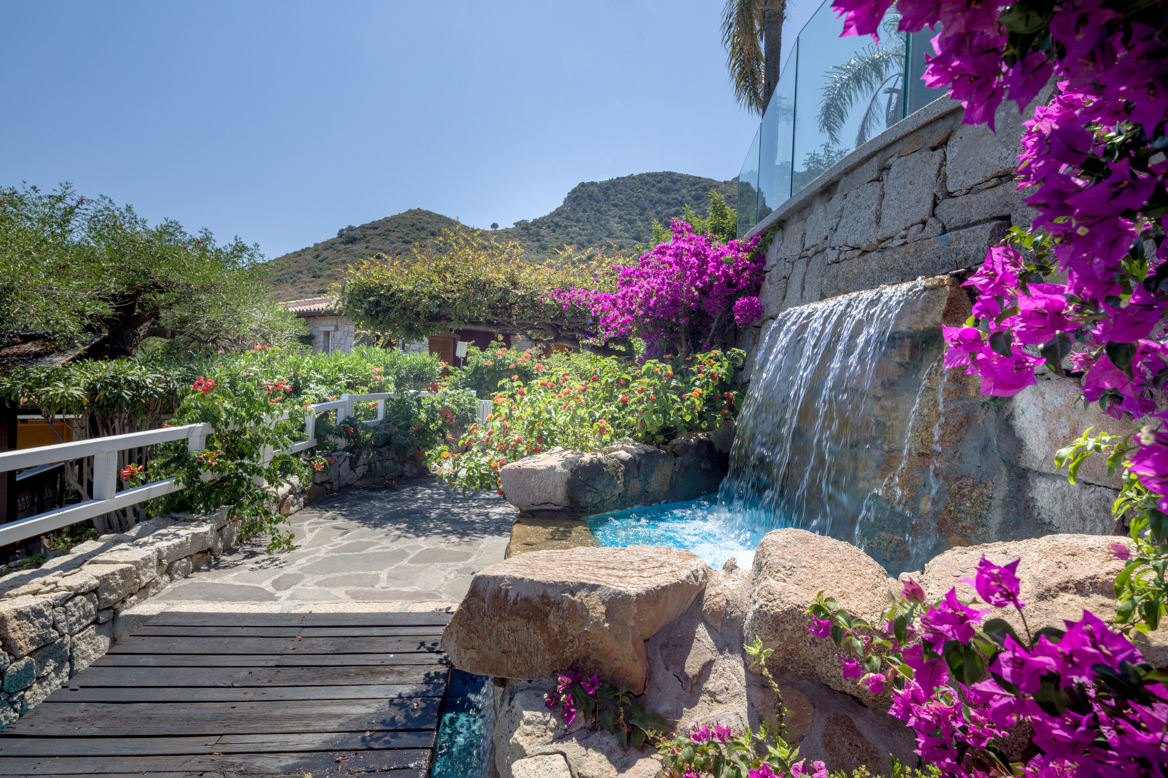 Ein Garten mit einer Holzterrasse, einem kleinen Wasserfall und leuchtend blühender Bougainvillea.