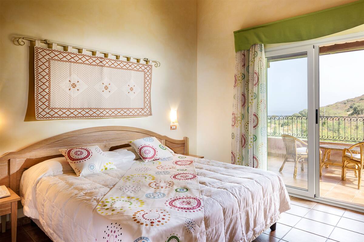 A bedroom with a bed, a nightstand, a wall-mounted decorative piece, and a sliding glass door leading to an outdoor area with a table and chairs.