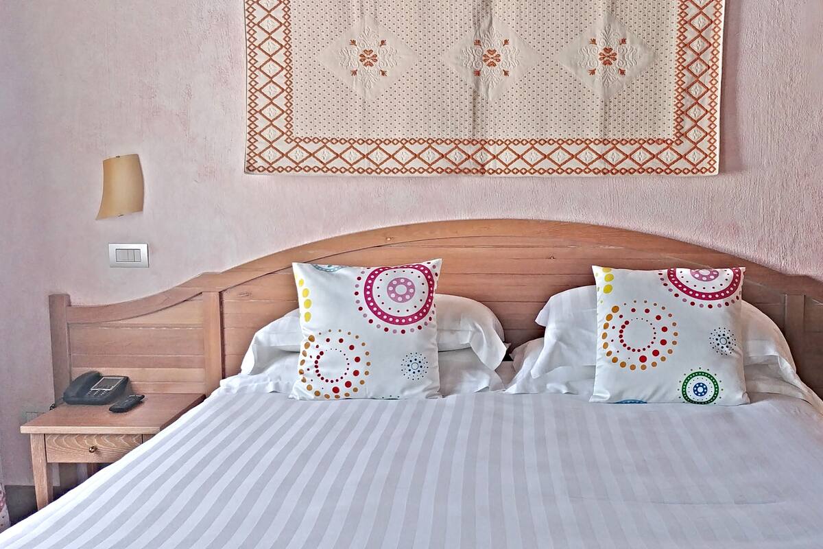 A neatly made bed with striped bedding, two pillows with colorful circular patterns, a wooden headboard, a bedside table with a telephone, and a wall-mounted decorative piece above the bed.