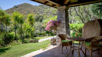 Une terrasse couverte, meublée de mobilier en osier, avec un pilier en pierre et une vue sur un jardin luxuriant et une colline.