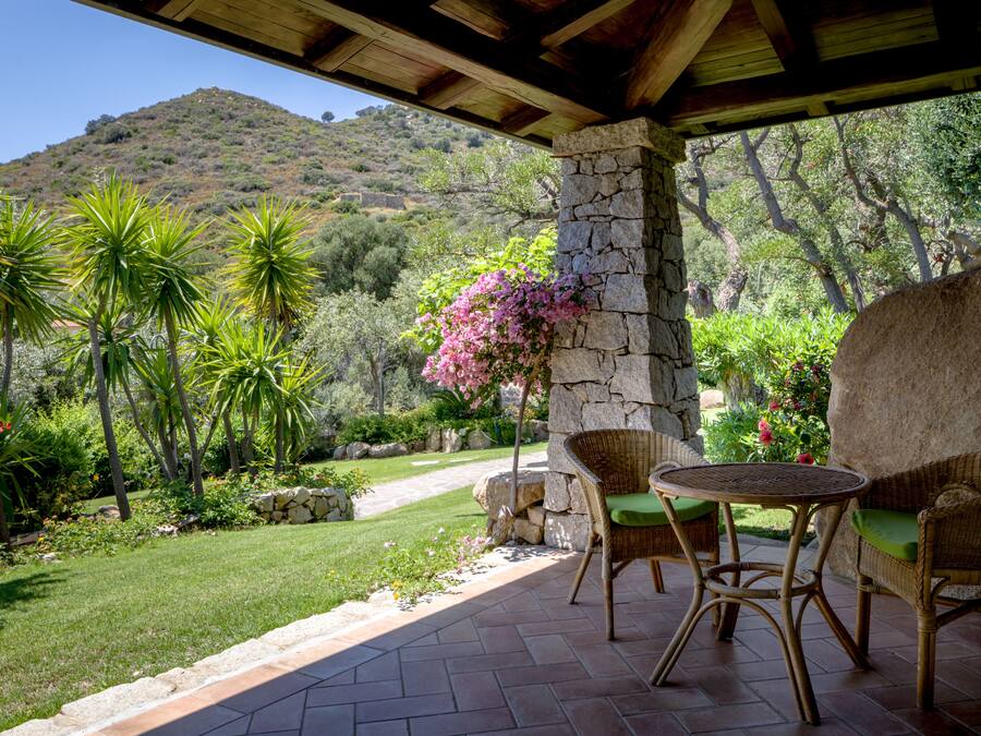 Une terrasse couverte, meublée de mobilier en osier, avec un pilier en pierre et une vue sur un jardin luxuriant et une colline.