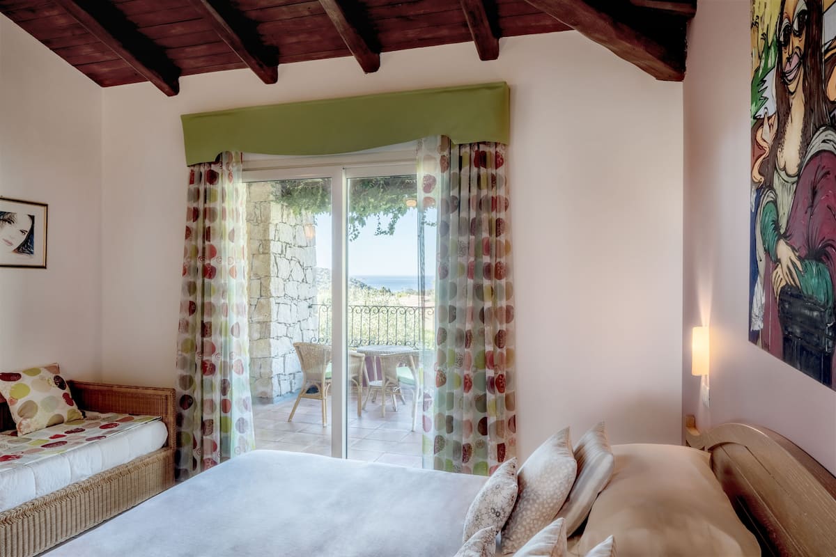 A bedroom with a bed, a wicker sofa, a painting, and a view of the outdoors.