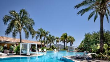Zona de piscina con sillones de descanso, palmeras y un edificio con tejado de tejas.