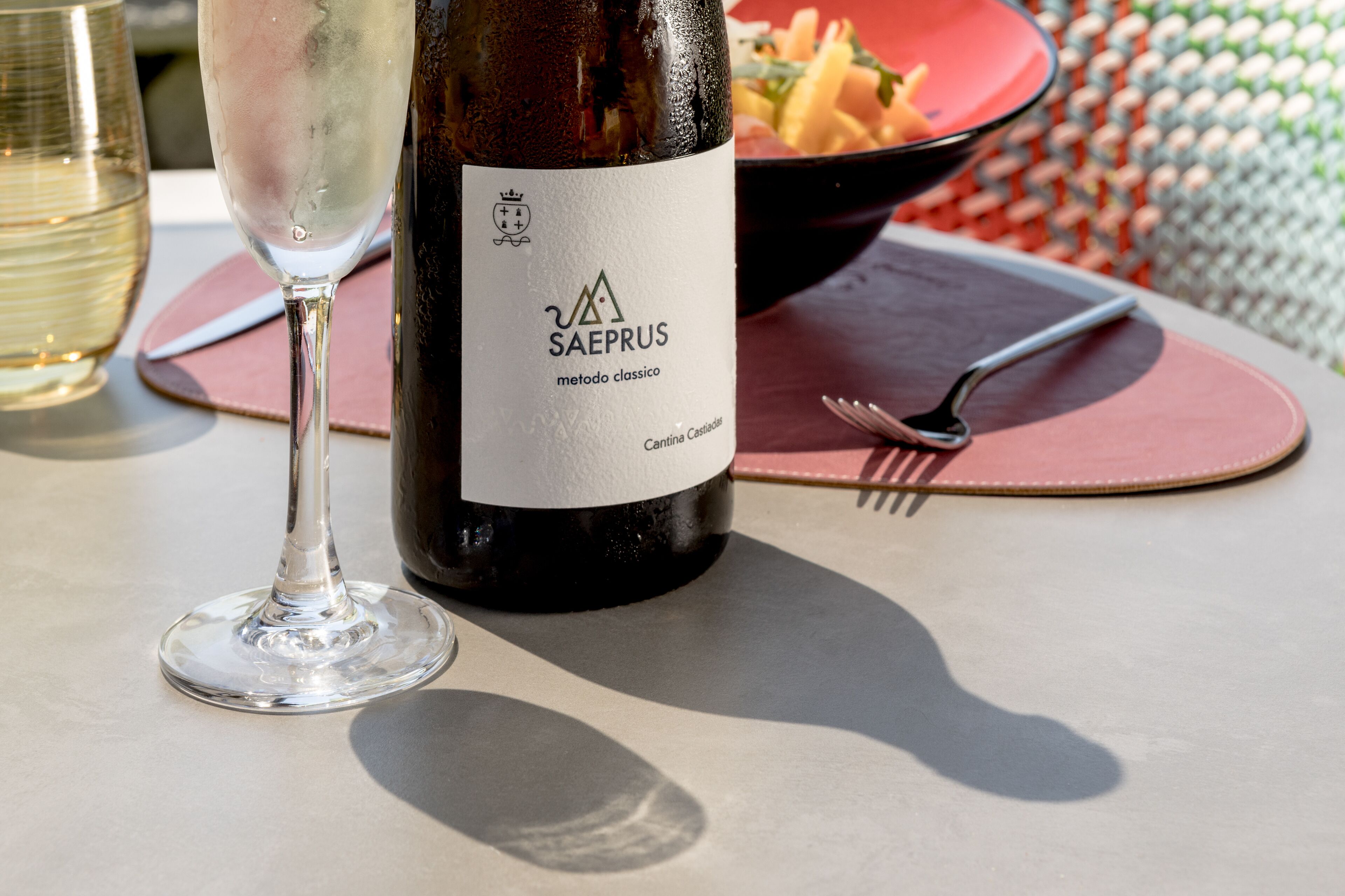 Uma garrafa de vinho com etiqueta, uma taça de vinho, uma tigela de comida e um garfo sobre uma mesa.