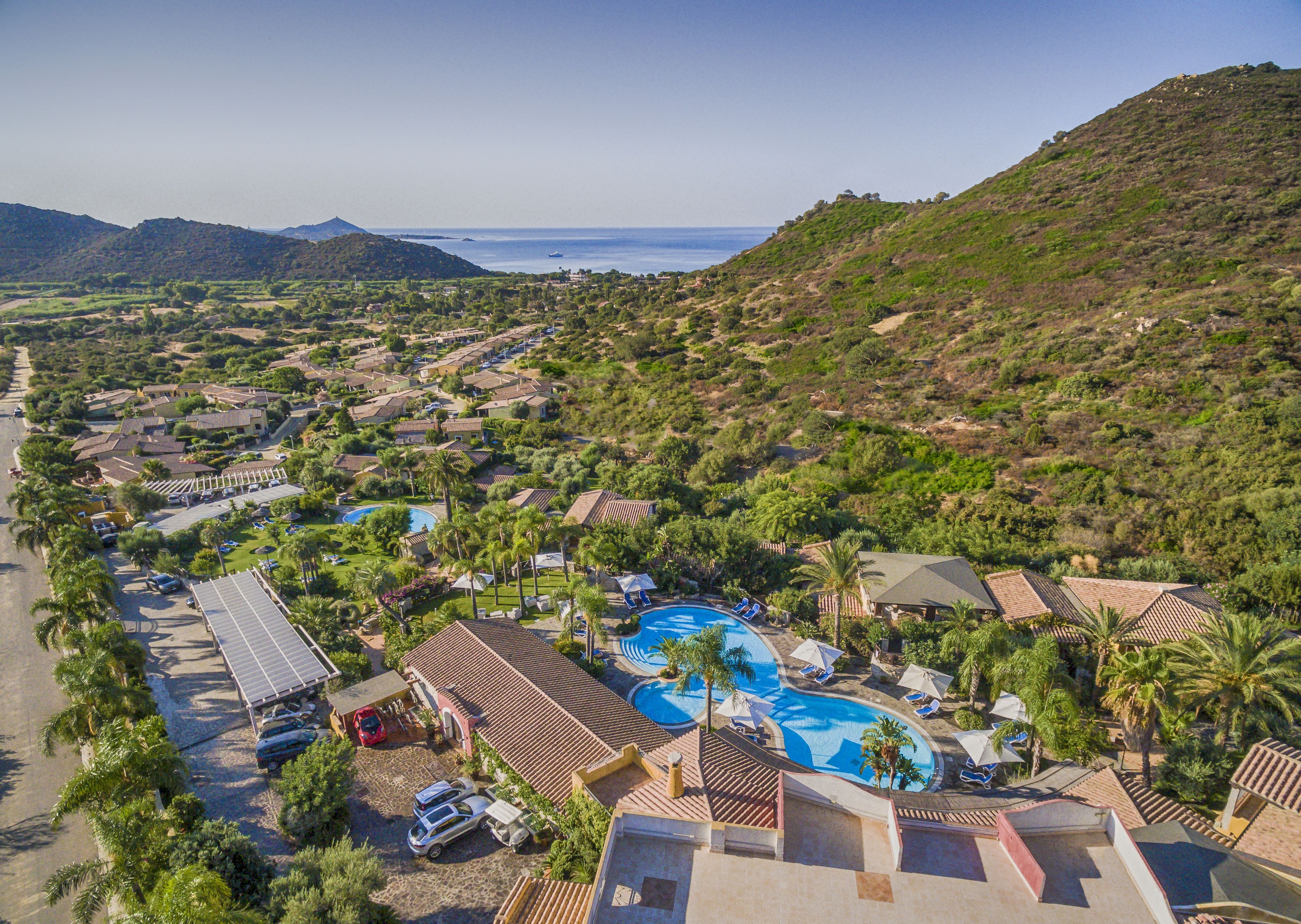 Vista aérea de un complejo turístico con varias edificaciones, piscina y vistas al mar y a las montañas.