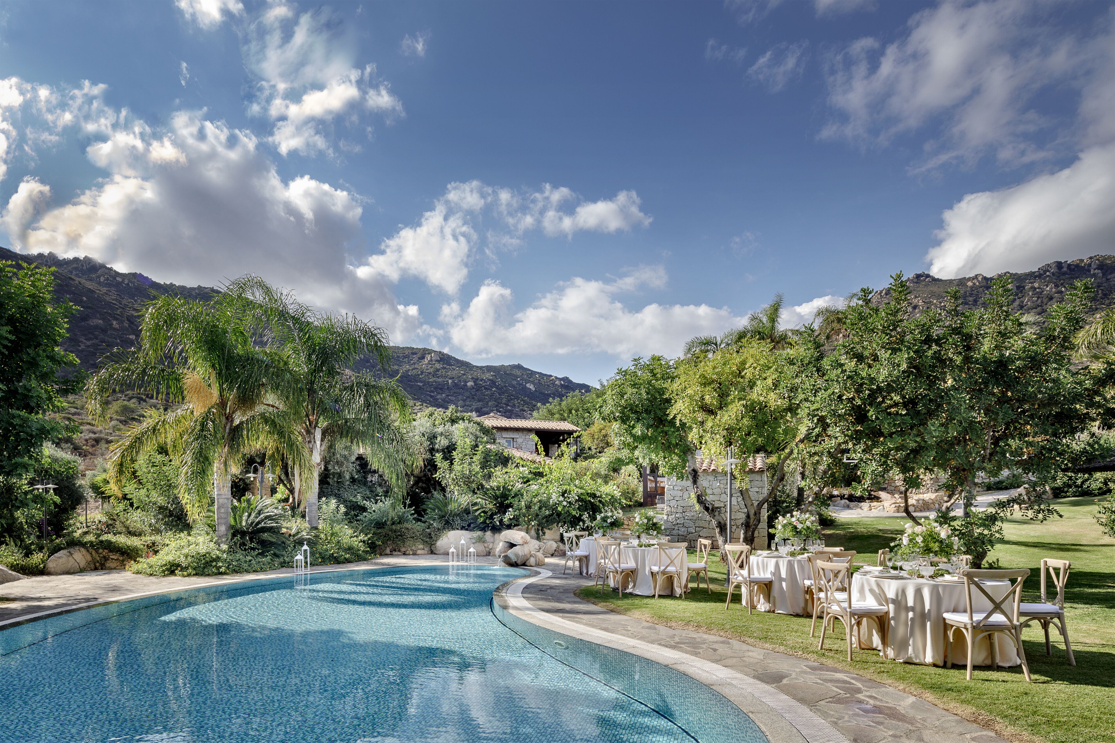 Uma área à beira da piscina com mesas e cadeiras, rodeada por vegetação exuberante e montanhas ao fundo.