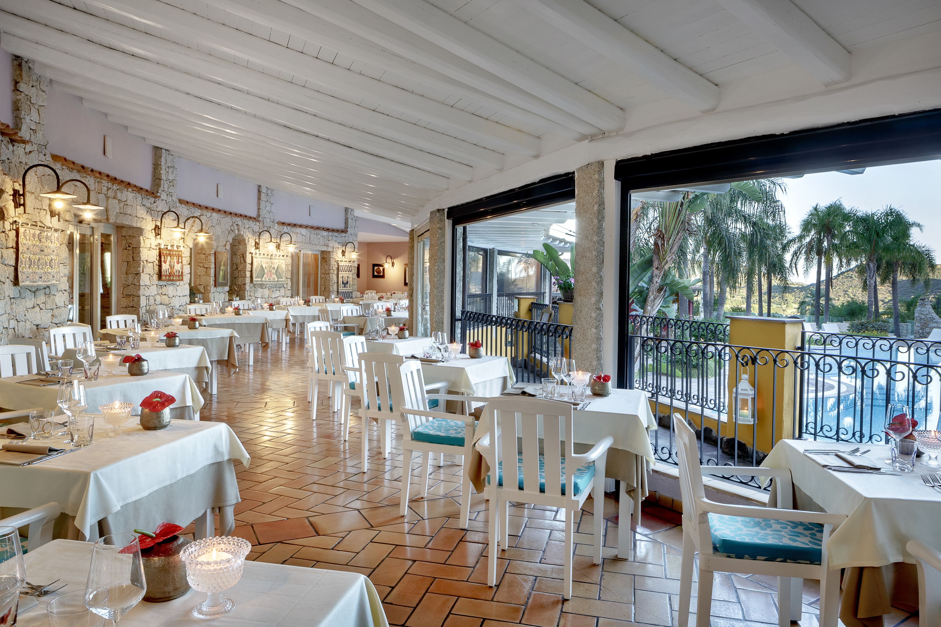 Restaurante com mesas e cadeiras brancas, paredes de pedra e vista para palmeiras.