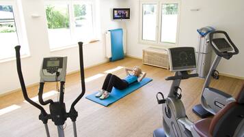 Une salle de sport équipée de matériel d’exercice, avec un téléviseur accroché au mur et de grandes fenêtres.
