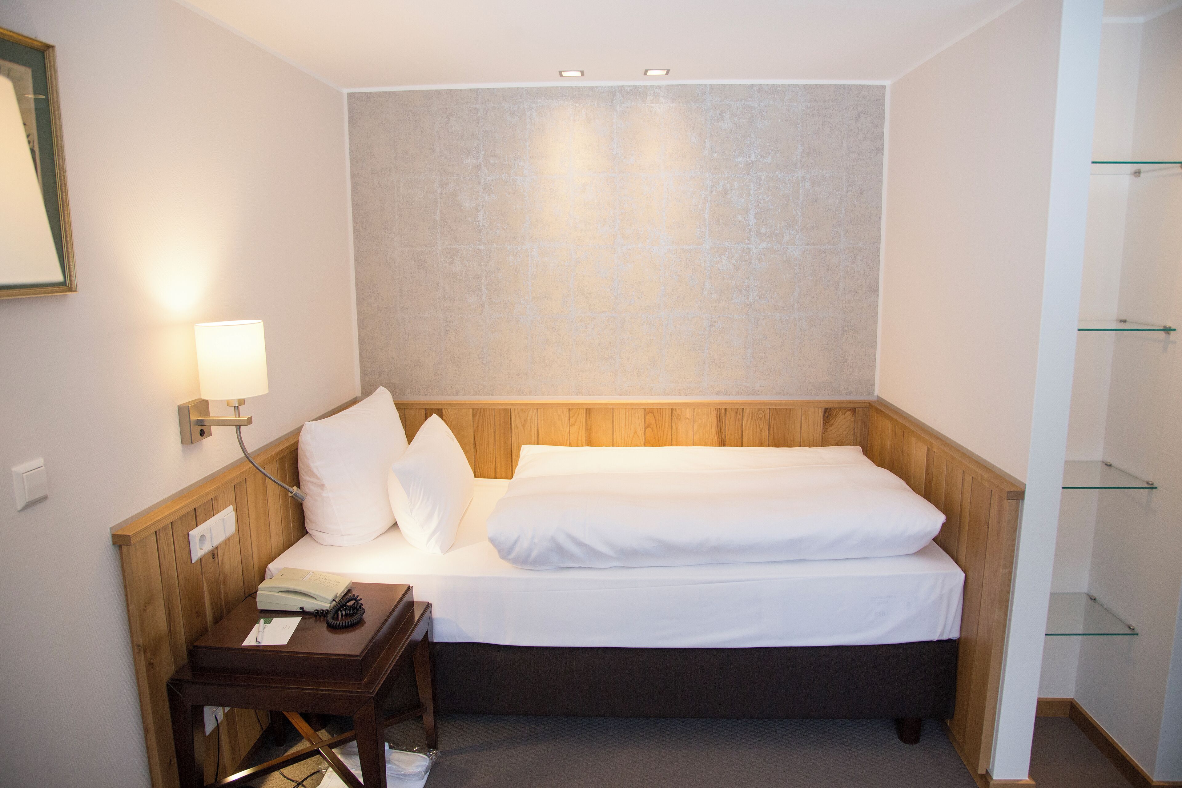 Ein kleines, ordentlich eingerichtetes Schlafzimmer mit einem Einzelbett, einem Nachttisch mit Lampe und einem an der Wand befestigten Regal.