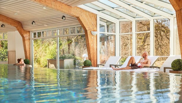 An indoor swimming pool with wooden beams, glass walls, and lounge chairs. - Hotel Jagdhaus Wiese (Schmallenberg)