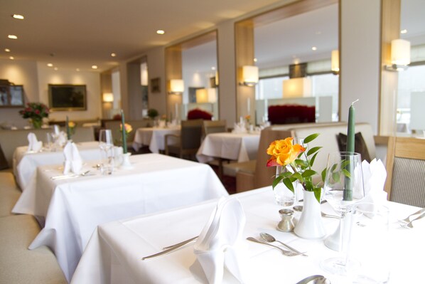 A restaurant with tables set for dining, featuring white tablecloths, silverware, and a vase with a flower.
