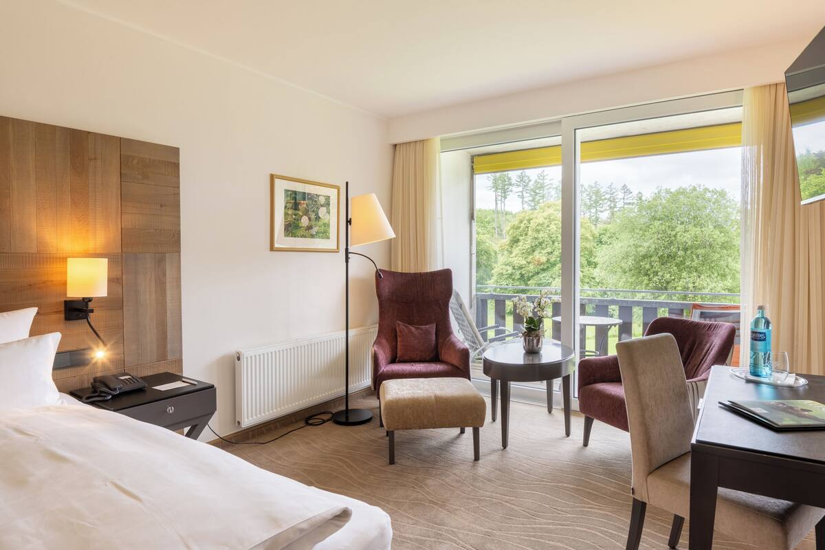 A hotel room with a bed, a desk, a chair, a small table, a lamp, and a window with a view of trees.