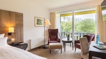 Habitación de hotel con cama, escritorio, silla, mesita, lámpara y ventana con vistas a árboles.