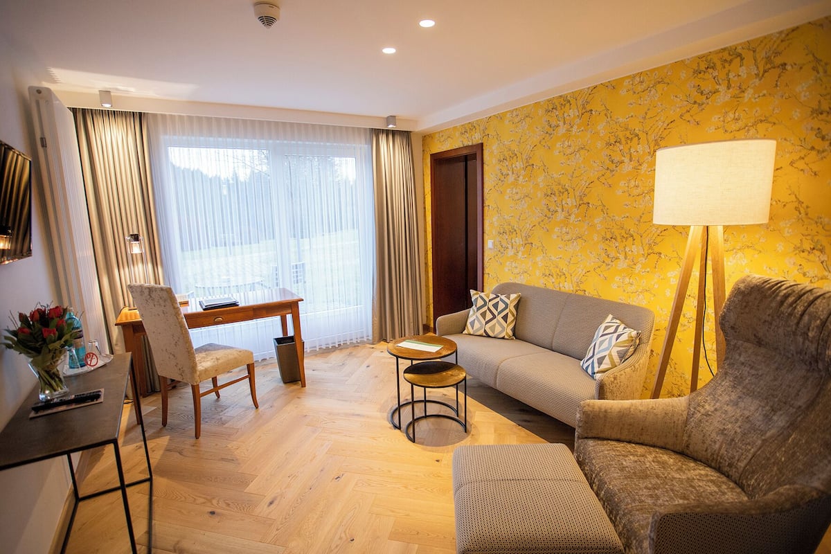 A modern living room with a sofa, armchair, and a small round table. There is a desk with a chair, a lamp, and a vase with flowers. The room has wooden flooring and a large window with curtains.