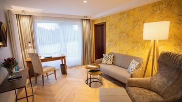 A modern living room with a sofa, armchair, and a small round table. There is a desk with a chair, a lamp, and a vase with flowers. The room has wooden flooring and a large window with curtains.