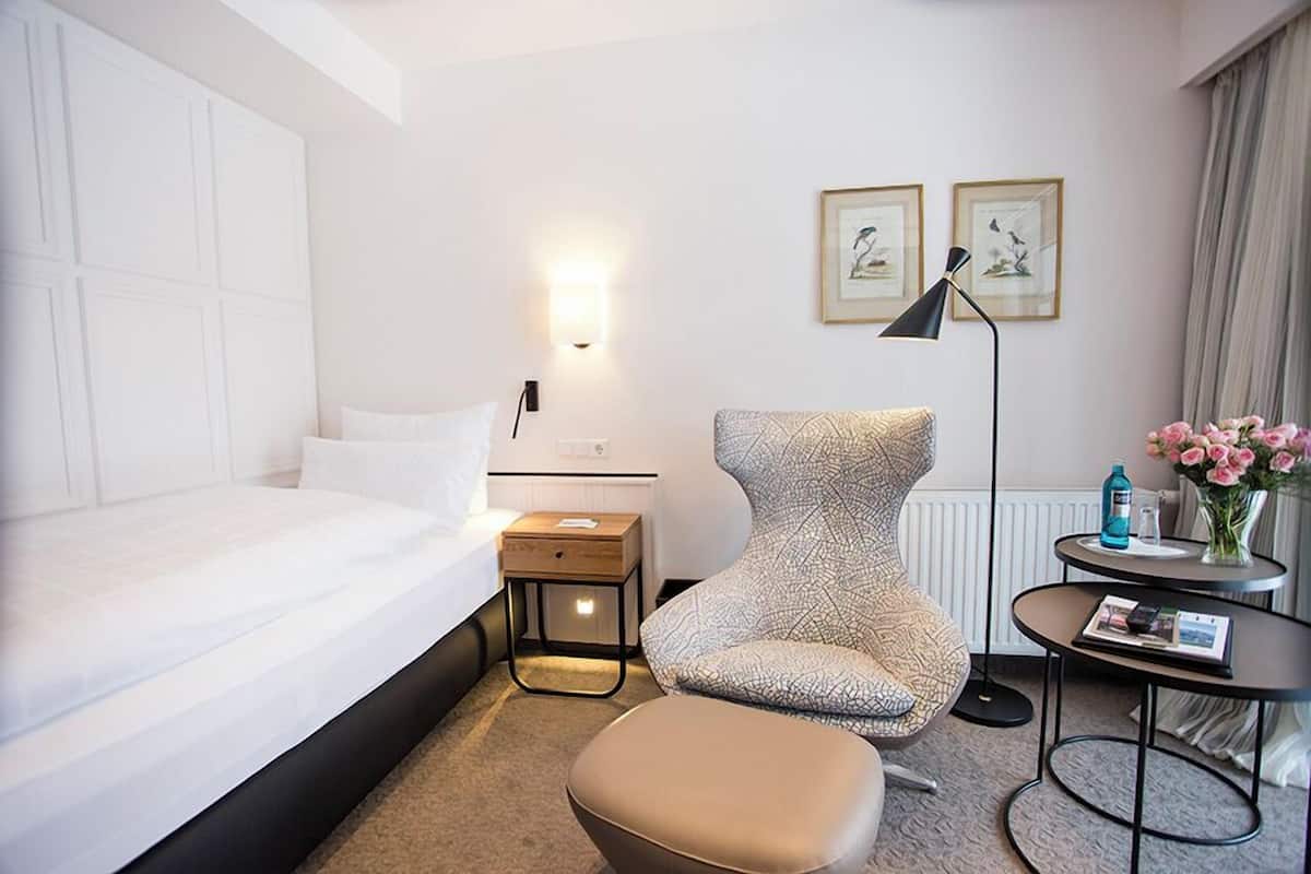 A hotel room with a bed, a chair, a small table with a lamp, and a vase with flowers.
