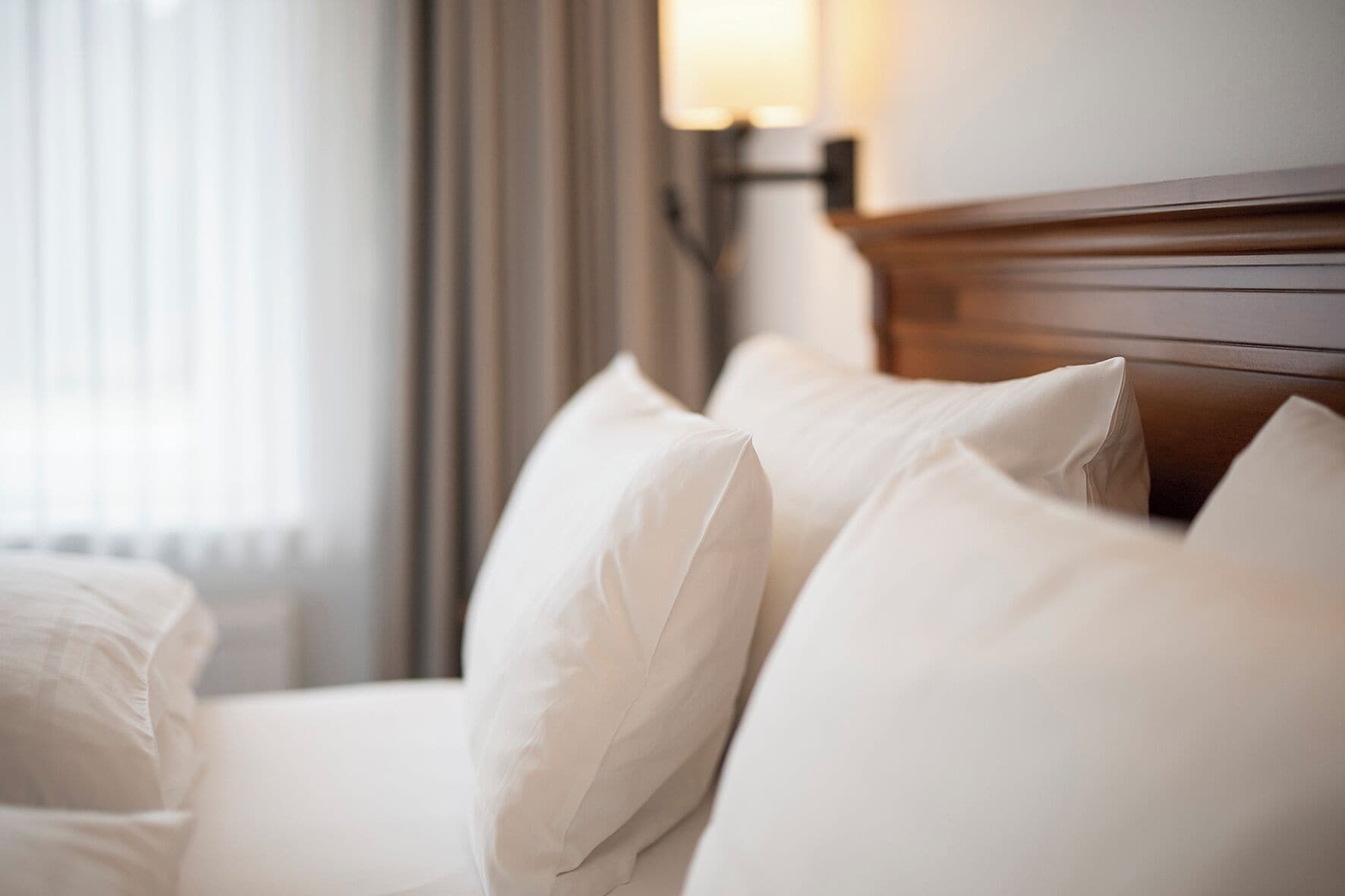 A neatly made bed with white linens and pillows, a wooden headboard, and a lamp on the nightstand.