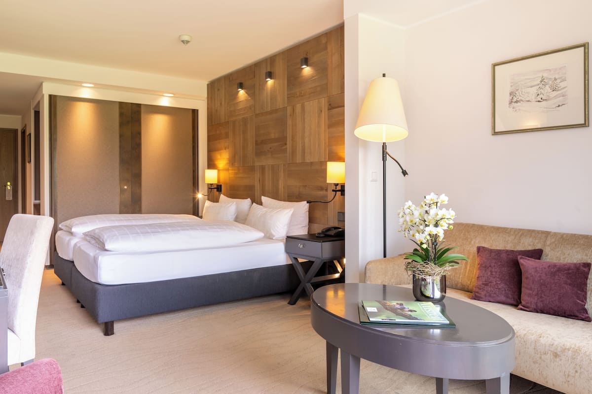 A modern hotel room with a large bed, a sofa, a small table with a lamp, and a potted plant.