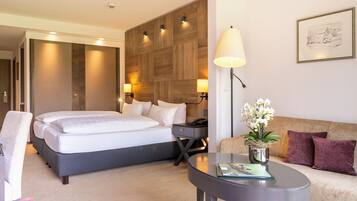 A modern hotel room with a large bed, a sofa, a small table with a lamp, and a potted plant.