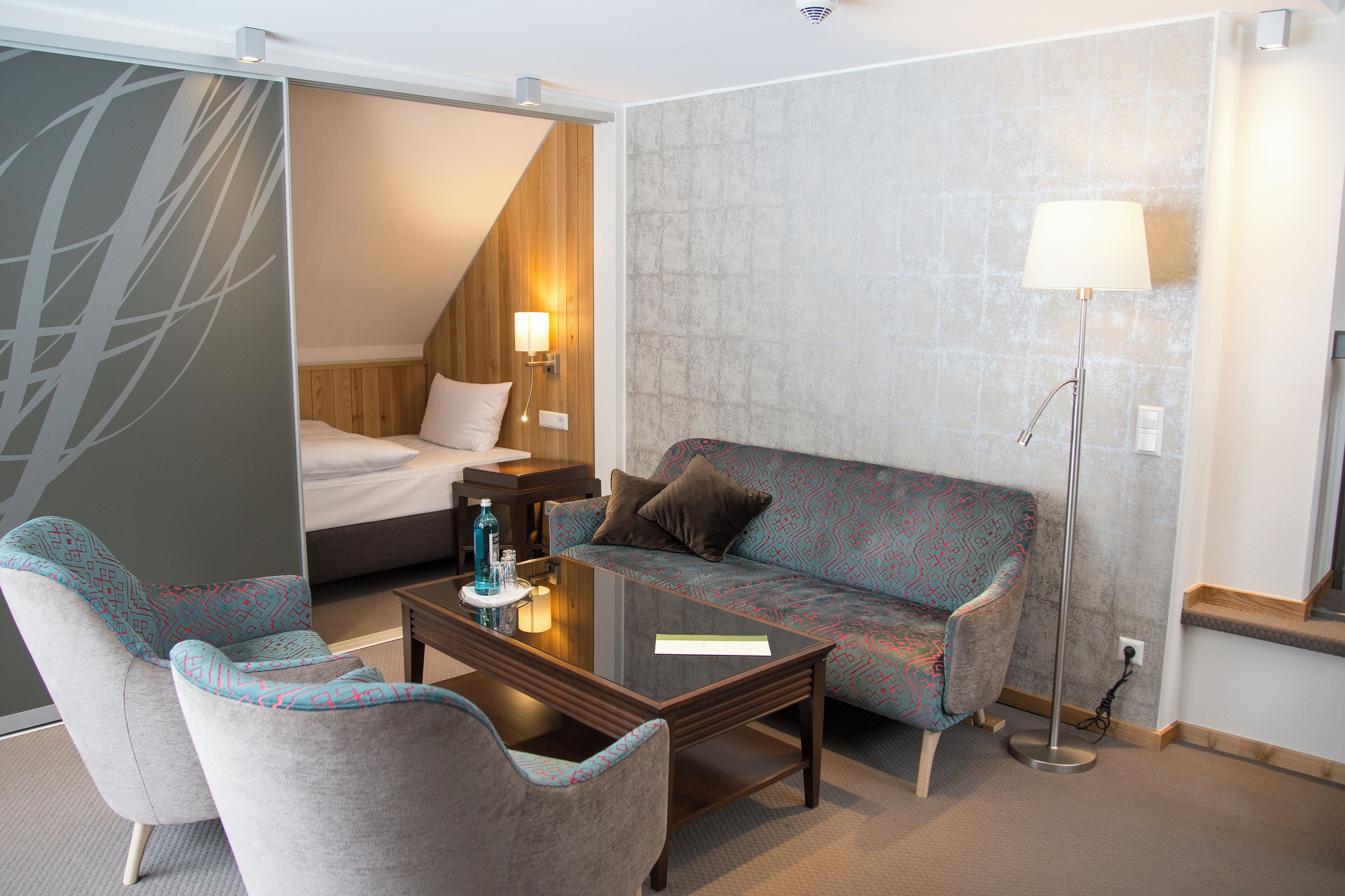 A hotel room with a bed, a sofa, two armchairs, a coffee table, and a floor lamp.