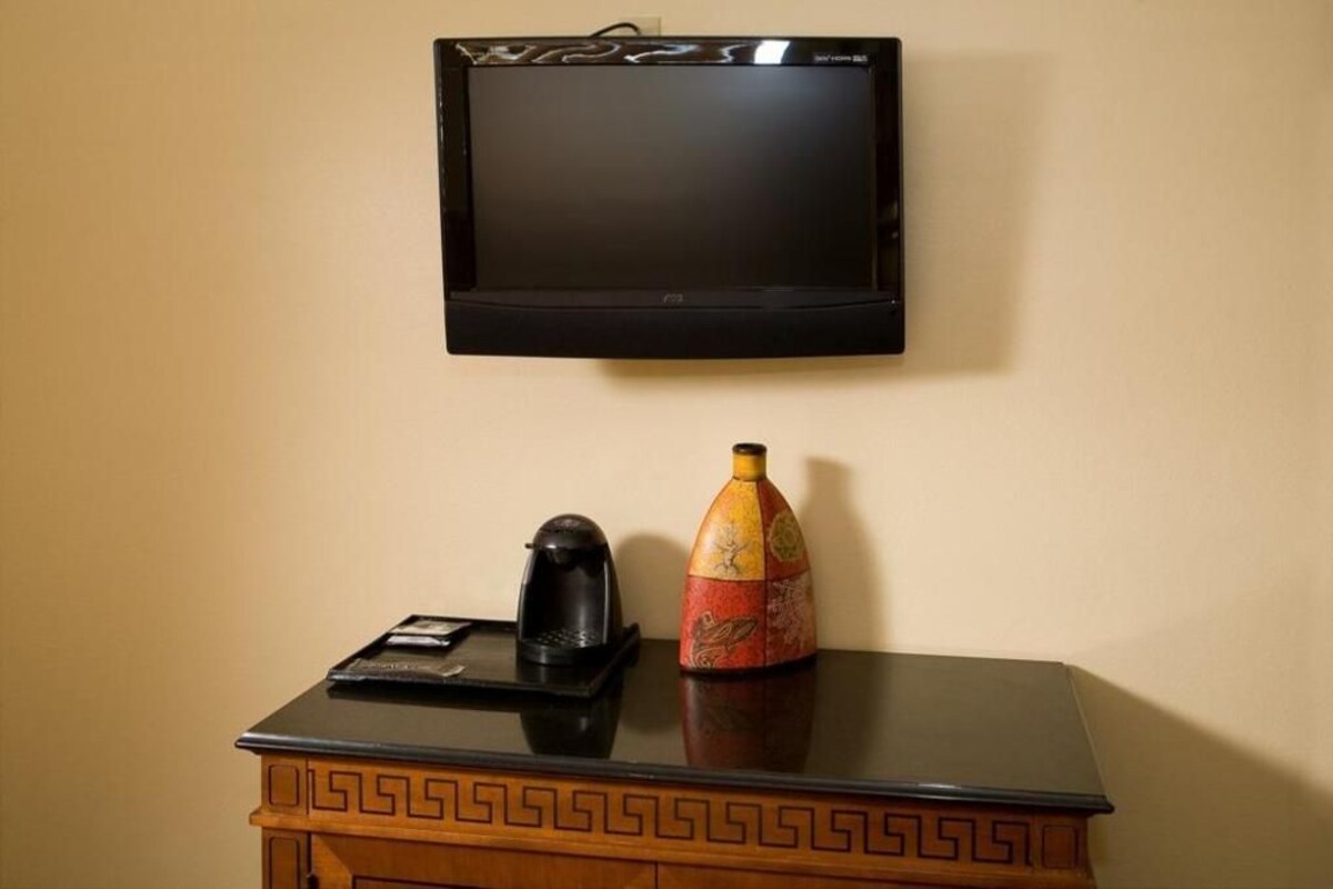 A television mounted on a wall above a wooden table with a decorative vase and a black object on it.