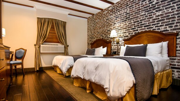A hotel room with two beds, a wooden headboard, a brick wall, a wooden floor, a desk with a chair, and a window with curtains. - Brand Plaza Hotel (Glendale)