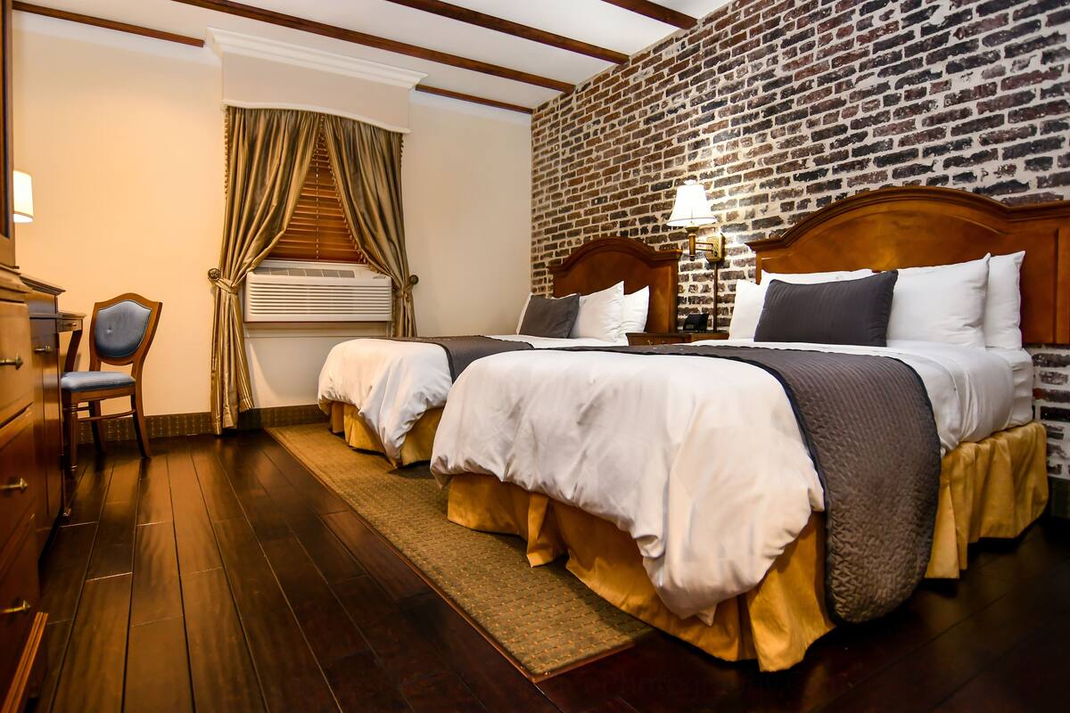 Habitación de hotel con dos camas, cabecero de madera, pared de ladrillo, piso de madera, escritorio con silla y ventana con cortinas.